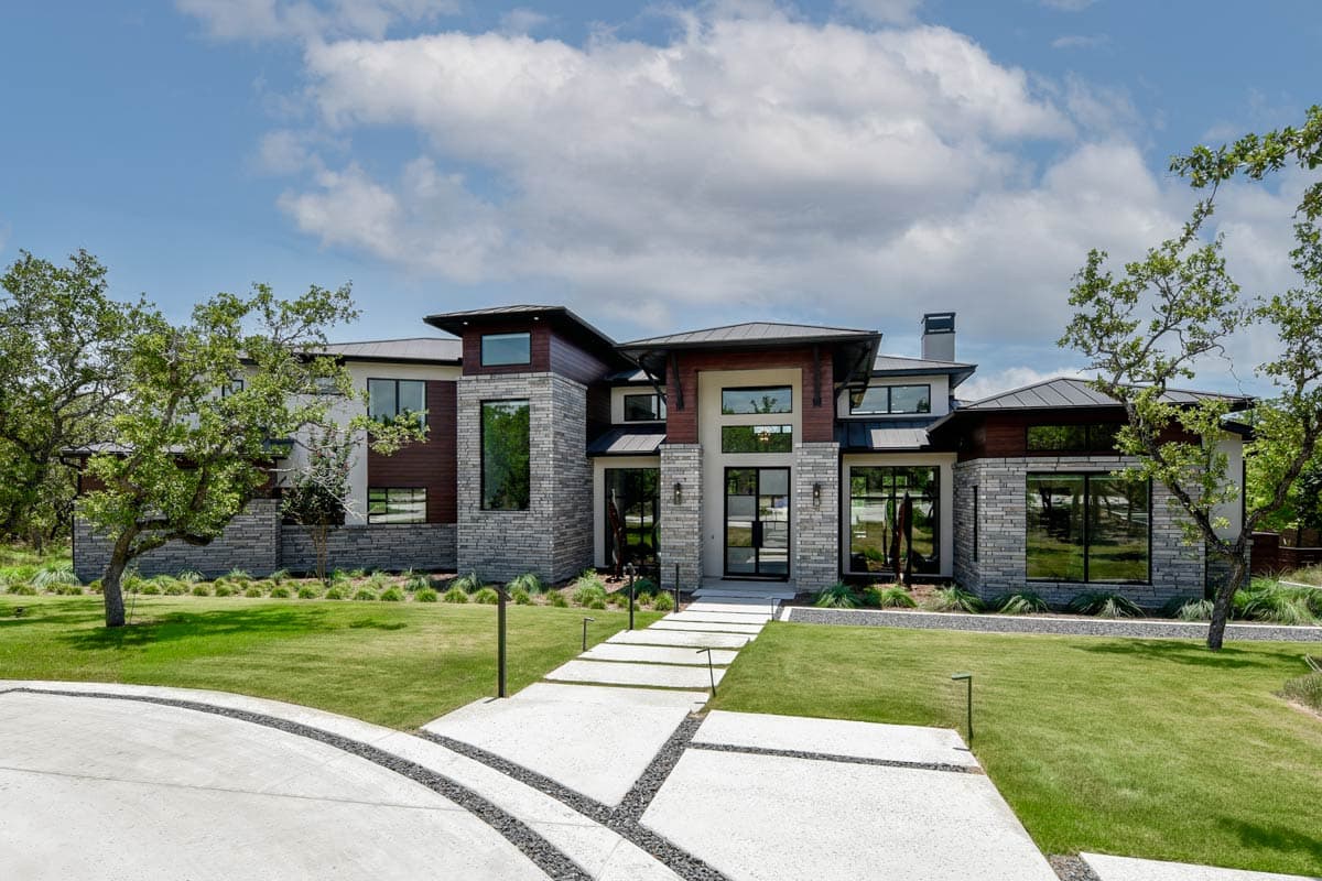 House plan exterior. Two-story modern house with stone facade, dark roof, large windows, and walkway to entryway with glass doors.