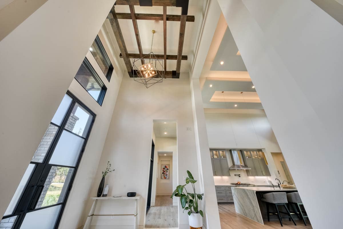 Interior view of a modern home with vaulted ceilings, exposed beams, geometric light fixture, large windows, and an open kitchen with island.