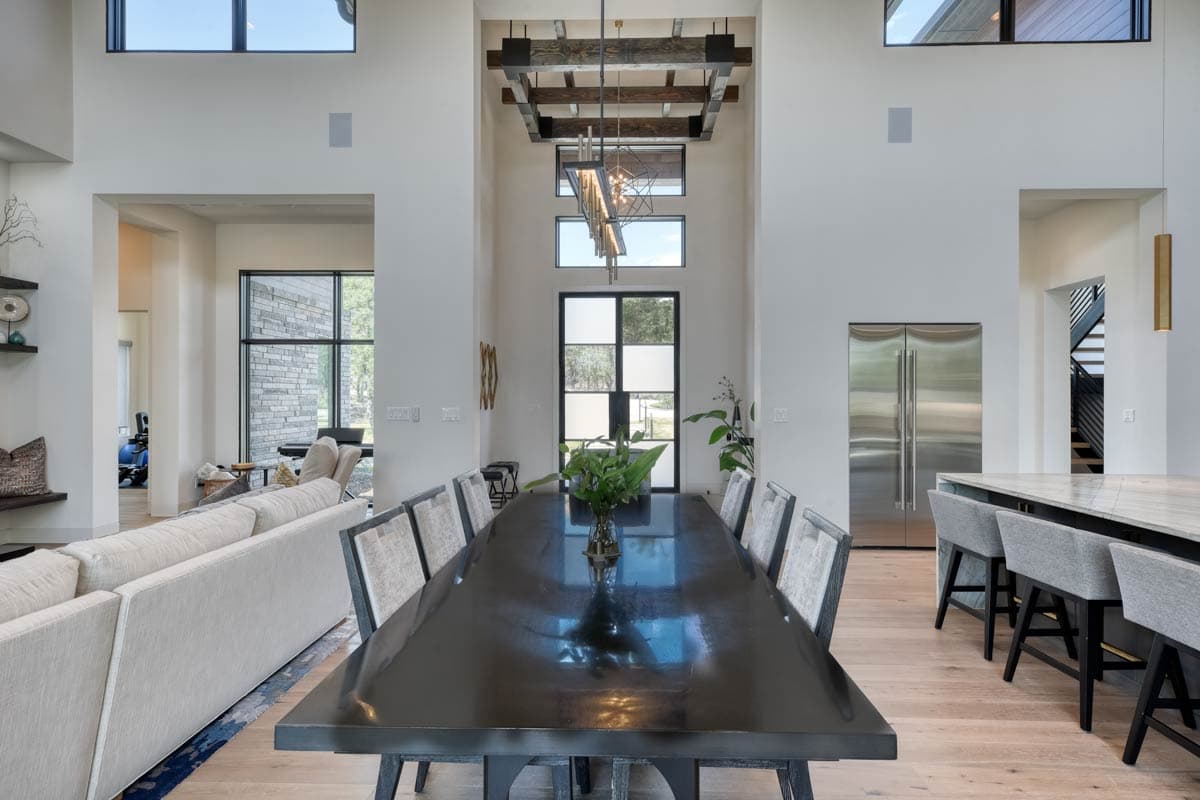 Great room with dining table, sectional sofa, kitchen island, and double refrigerators. Features vaulted ceiling with exposed beams and large windows.