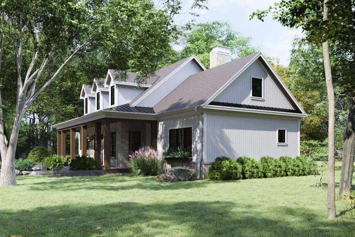 A white farmhouse with a stone porch and dark roof, nestled among trees. The yard is grassy, with trimmed bushes and decorative rocks.