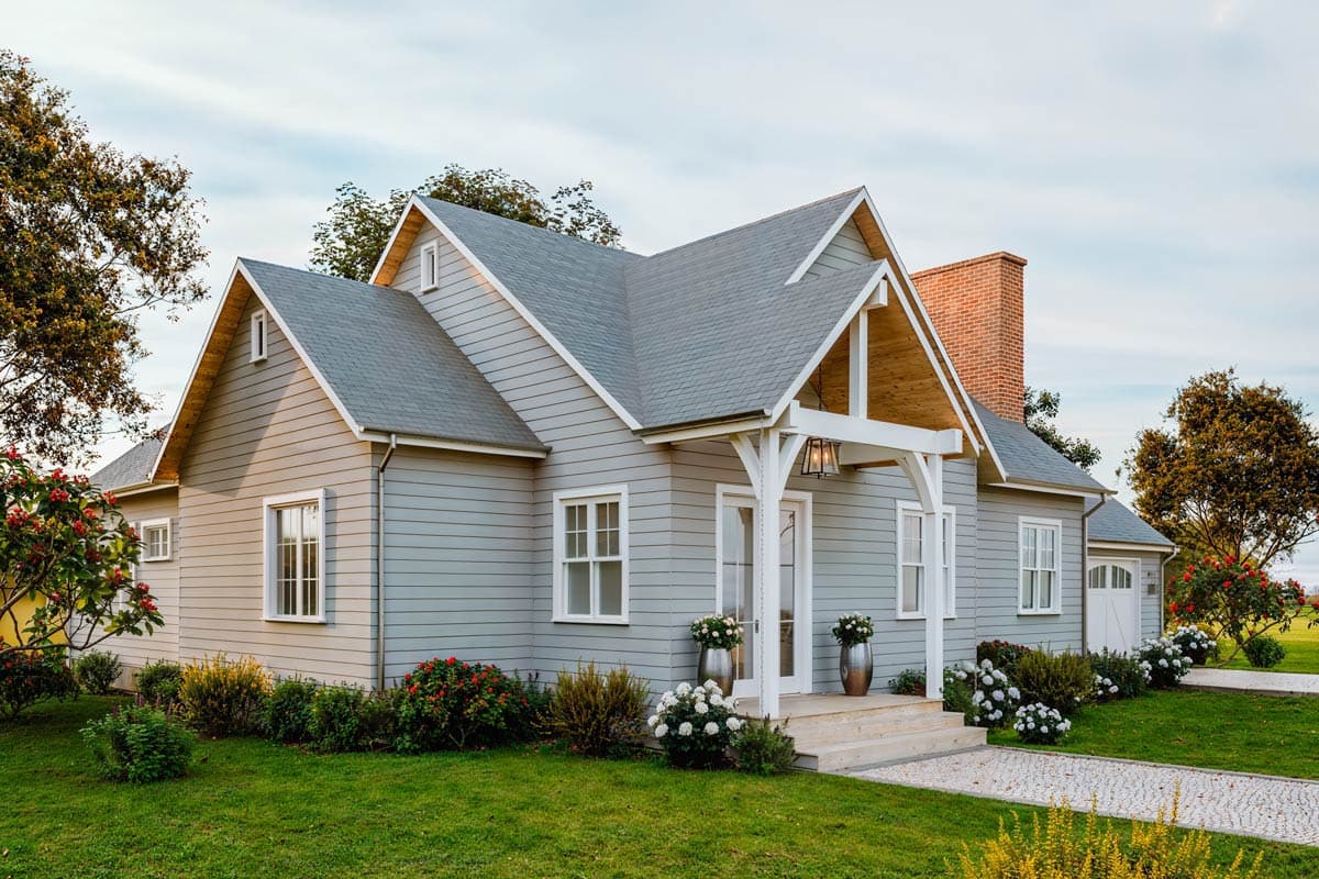 A charming grey house with a white porch and steps, a brick chimney, and well-kept landscaping under a clear sky.