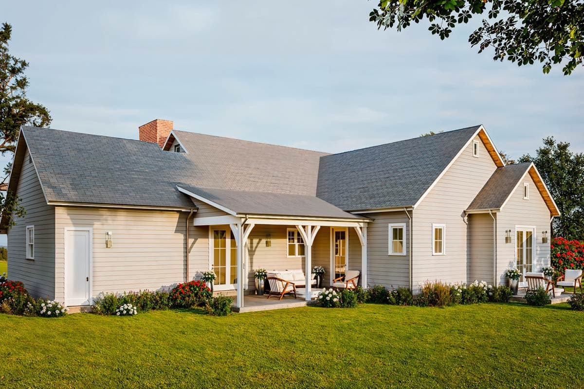 Exterior shot of a modern farmhouse with gray siding, a porch, and a well-manicured lawn.  Sunny day.