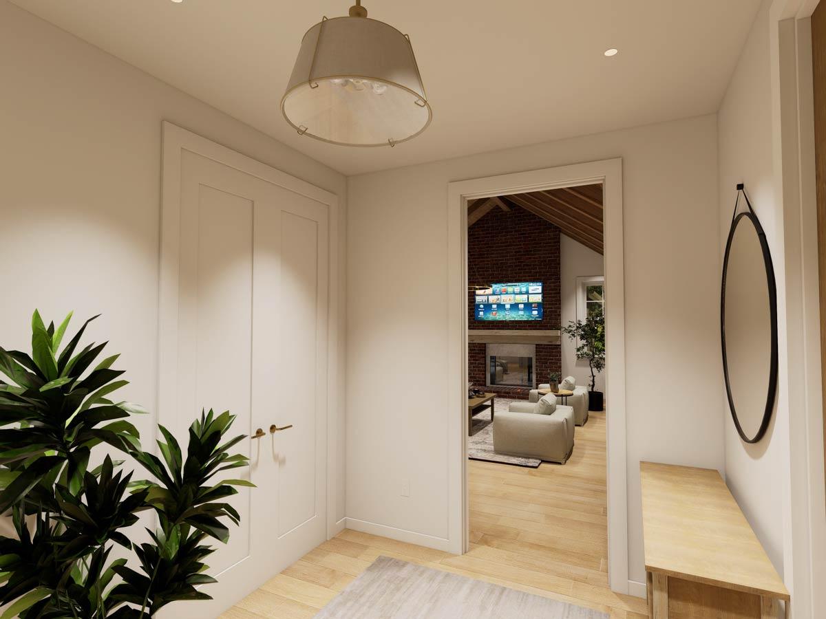 Interior view of a hallway showing a door, large plant, and opening to a great room with a vaulted ceiling and brick fireplace.