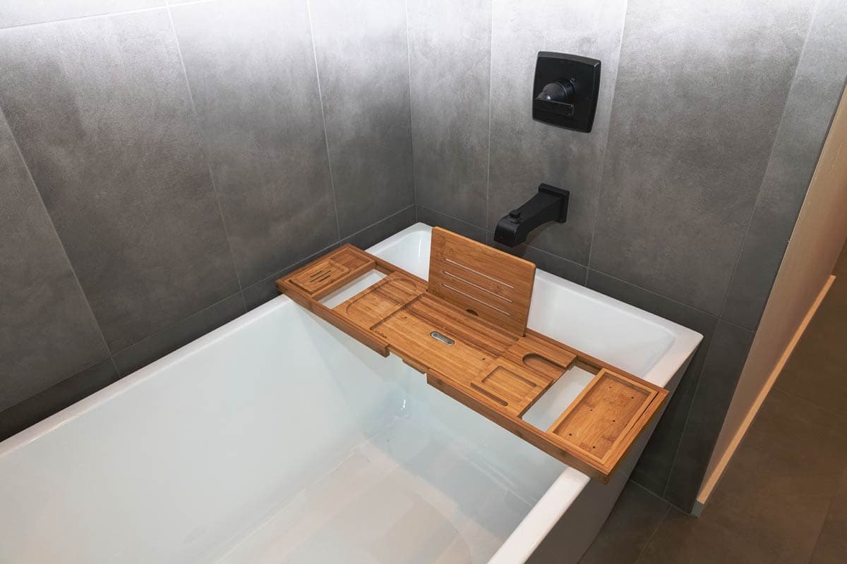 Bathtub with a wooden bath caddy, black faucet, and gray tile walls.