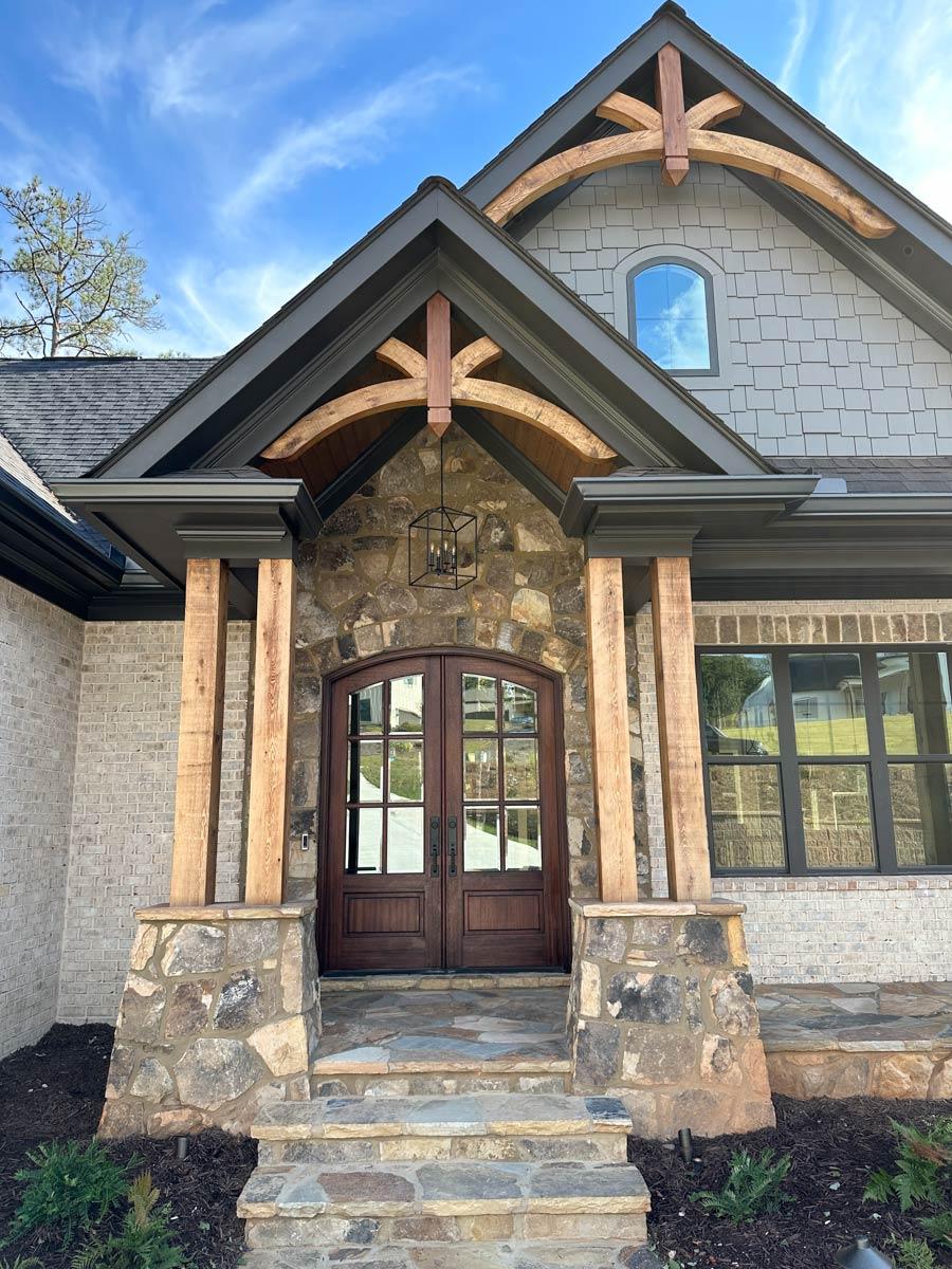House plan exterior with stone base, wooden posts, arched entry, double doors, gable roof, and decorative timber framing.
