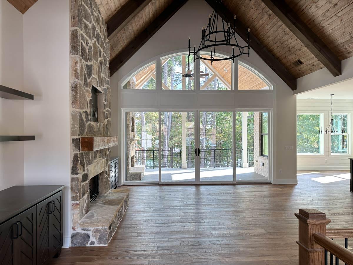Great room with stone fireplace, vaulted wood-beamed ceiling, and large arched windows overlooking trees.