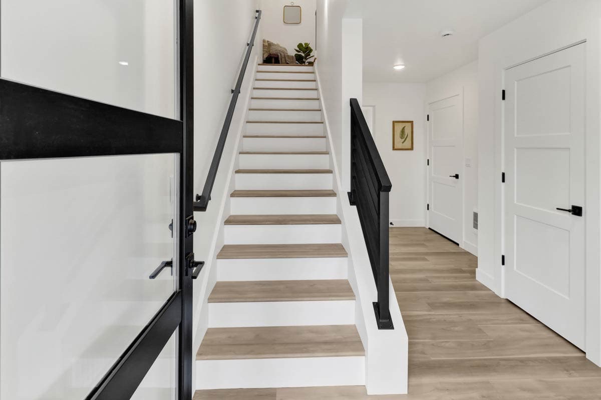 Interior view of a staircase with white risers, wood treads, and black railings. Doors and wood-look flooring visible.