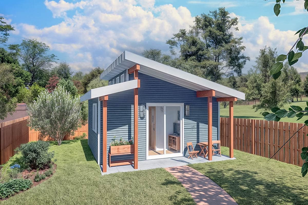 A small, modern blue house with a porch and garden, set against a backdrop of trees under a partly cloudy sky.