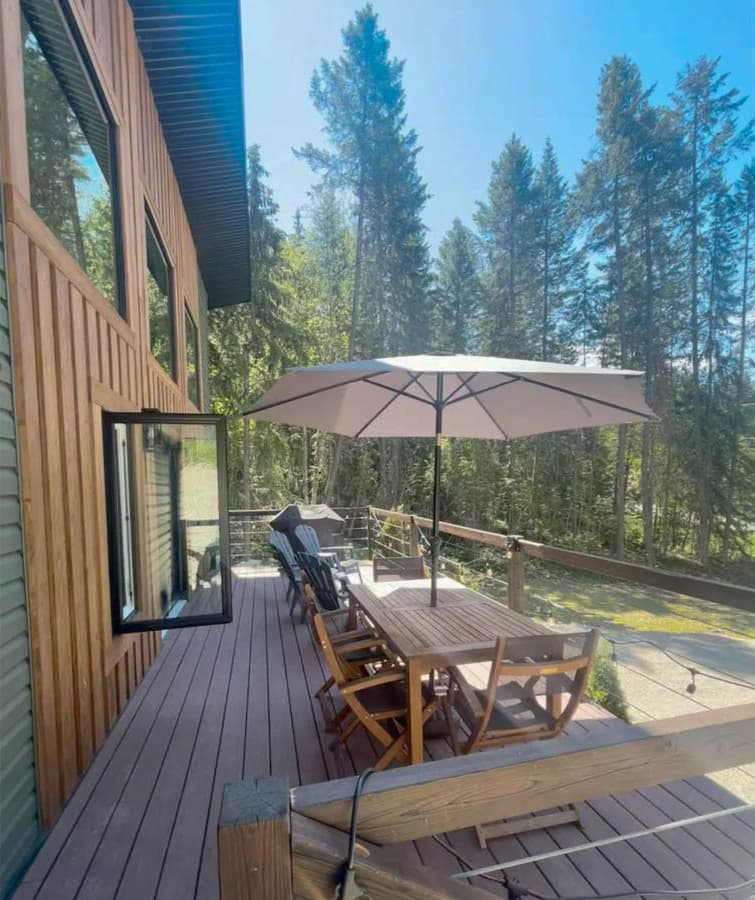 Modern house exterior with wood siding, large windows, and expansive deck featuring outdoor dining set and umbrella.