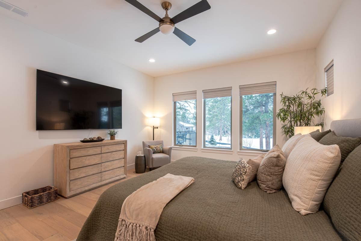 Primary bedroom with king bed, wall-mounted TV, dresser, ceiling fan, and three large windows overlooking a wooded area.