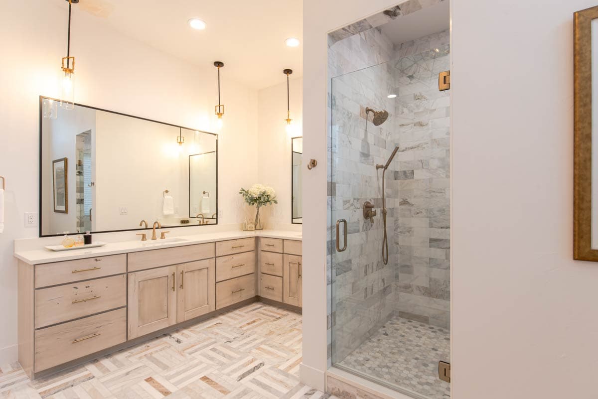 Bathroom with a dual vanity featuring light wood cabinets and a frameless glass shower with marble tile walls and brass fixtures.