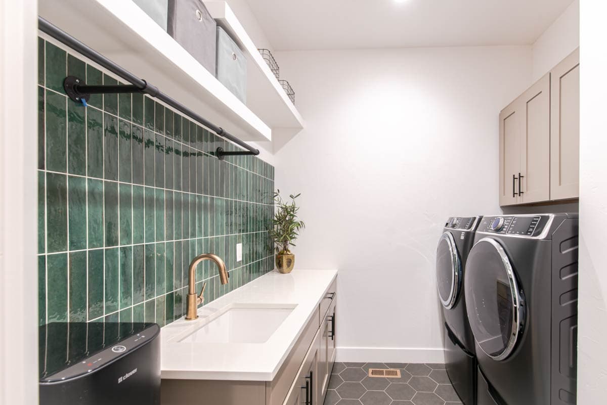 Laundry room with stacked washer/dryer, sink, countertop, green vertical tile backsplash, and upper cabinets.