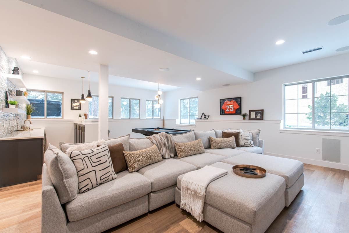 Modern basement living room with sectional sofa, pool table, and wet bar. Large windows offer natural light.