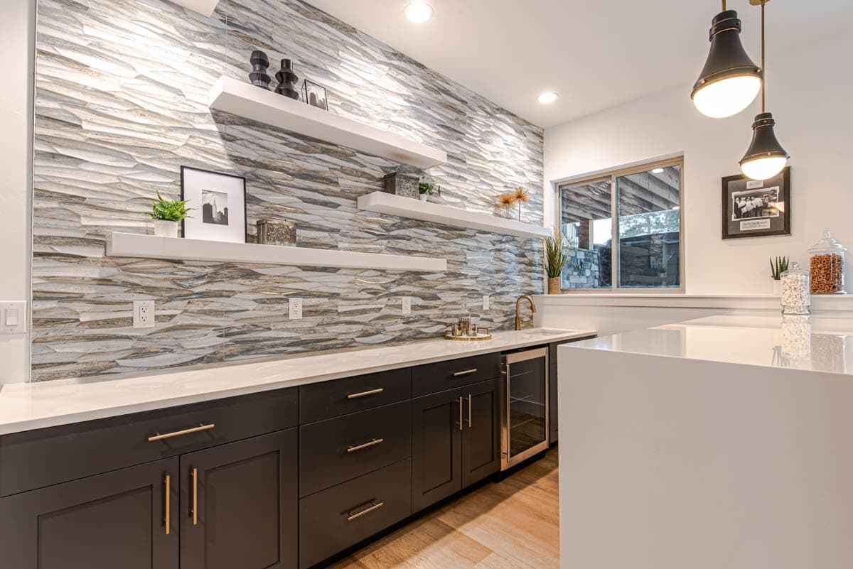 Bar area with a textured stone accent wall, white floating shelves, dark cabinetry, wine fridge, and a white countertop.