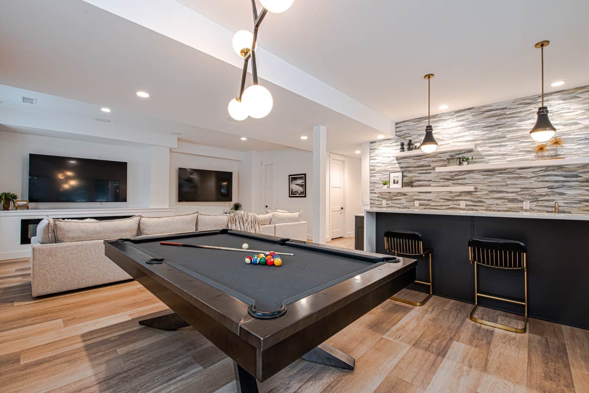 Finished basement with pool table, sectional sofa, large screen TVs, and a bar area with stone accent wall and pendant lighting.