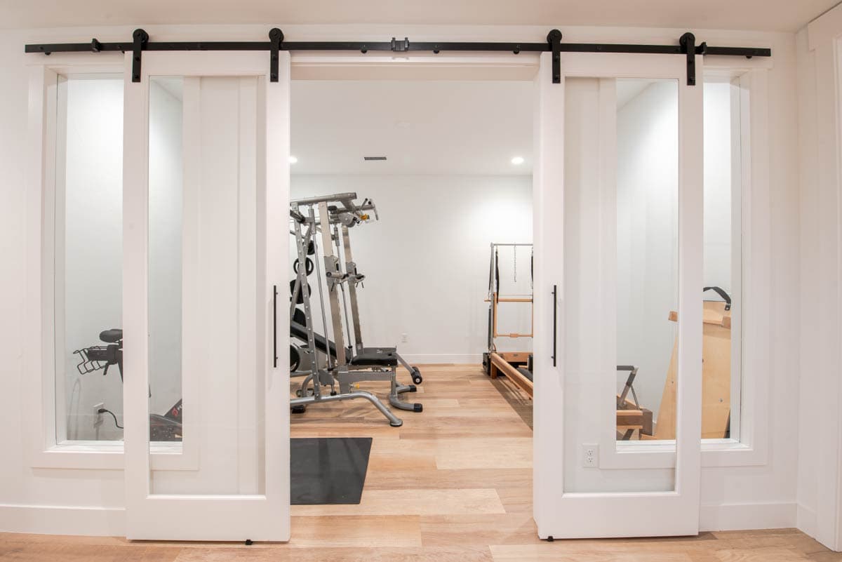 Interior view of a home gym featuring a weight machine, Peloton bike, and Pilates reformer, accessible via white sliding barn doors with glass panels.