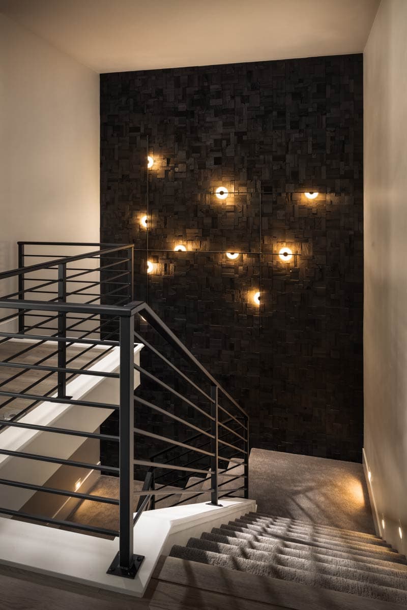 Staircase with modern metal railings ascends past a dark wood paneled accent wall with scattered round sconces.