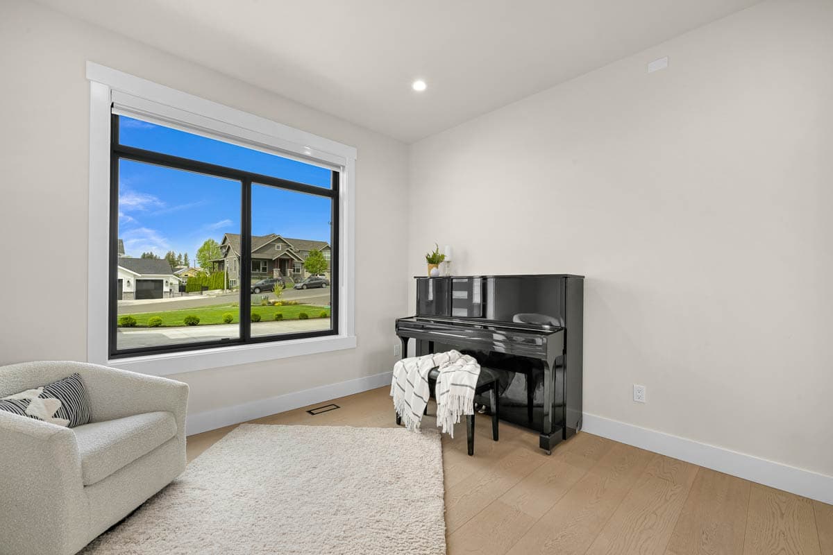 Interior view of a room with a piano, armchair, large window overlooking a street with houses, and light wood floors.
