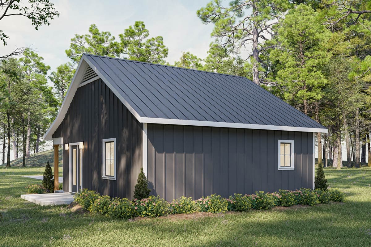 A modern, gray-sided cottage with a dark metal roof sits nestled in a grassy clearing surrounded by lush green trees on a sunny day.