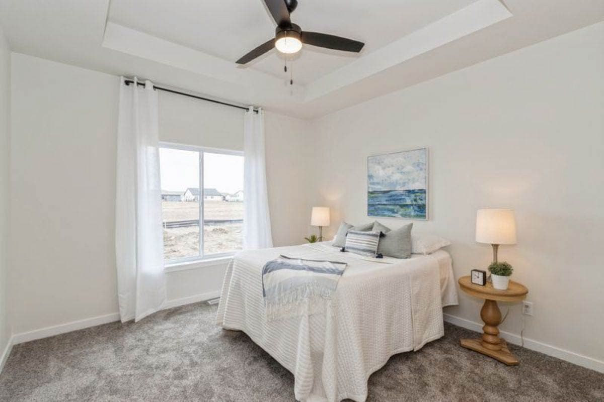 Bedroom with tray ceiling, ceiling fan, large window with white curtains, and a bed with white bedding.