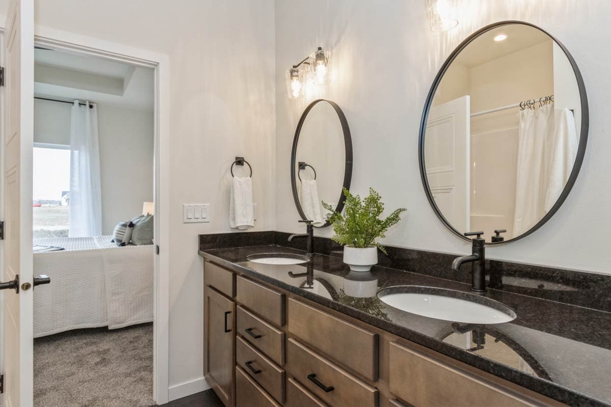 Bathroom vanity with double sinks, granite countertop, oval mirrors, and doorway to bedroom.