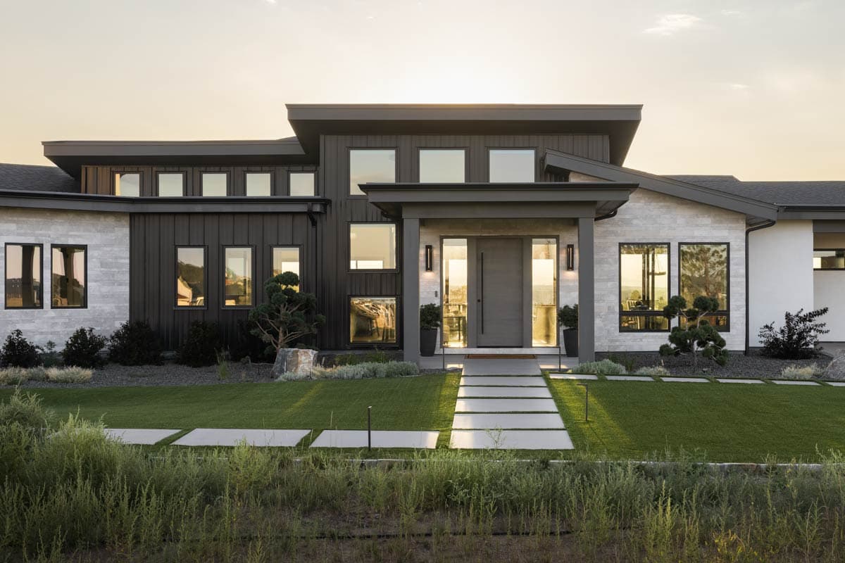 Modern house plan exterior with a flat roof, dark metal siding, stone accents, and a prominent covered entryway with large glass panels.
