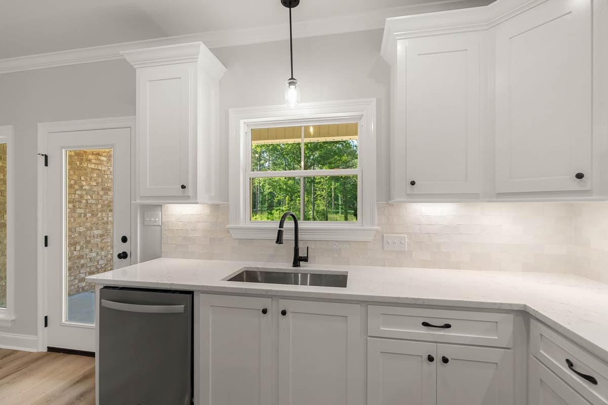 Kitchen interior with white shaker cabinets, quartz countertops, stainless steel sink and dishwasher, and a double-hung window.