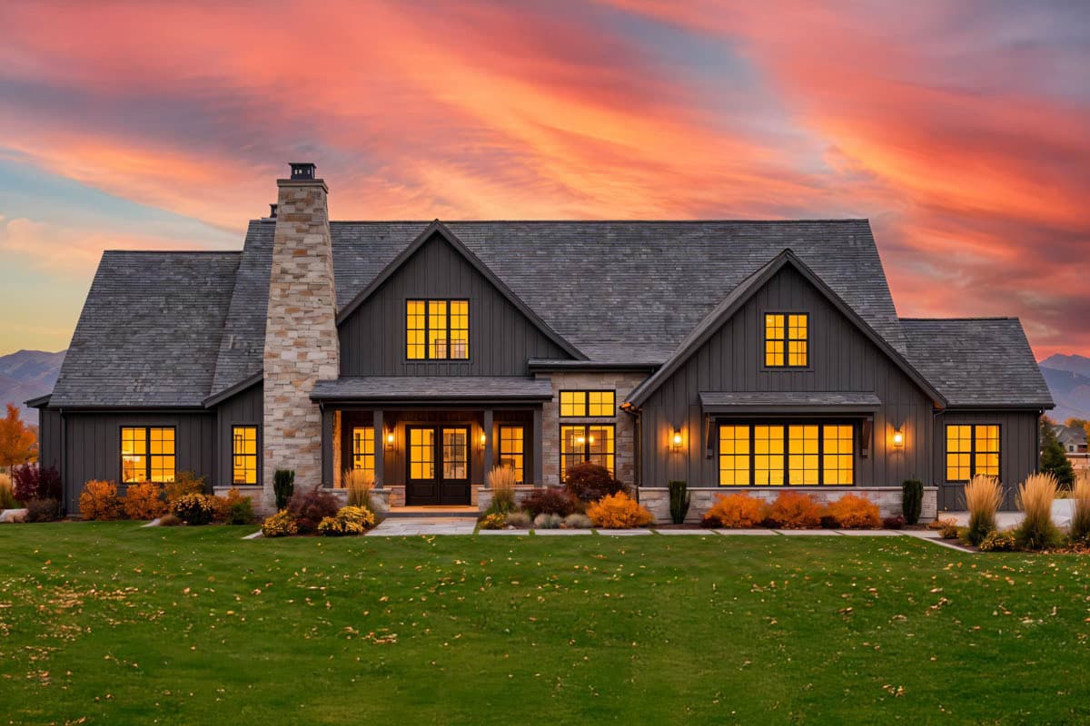 A large, modern farmhouse with dark siding and a stone chimney stands against a vibrant sunset. Glowing windows illuminate the interior.