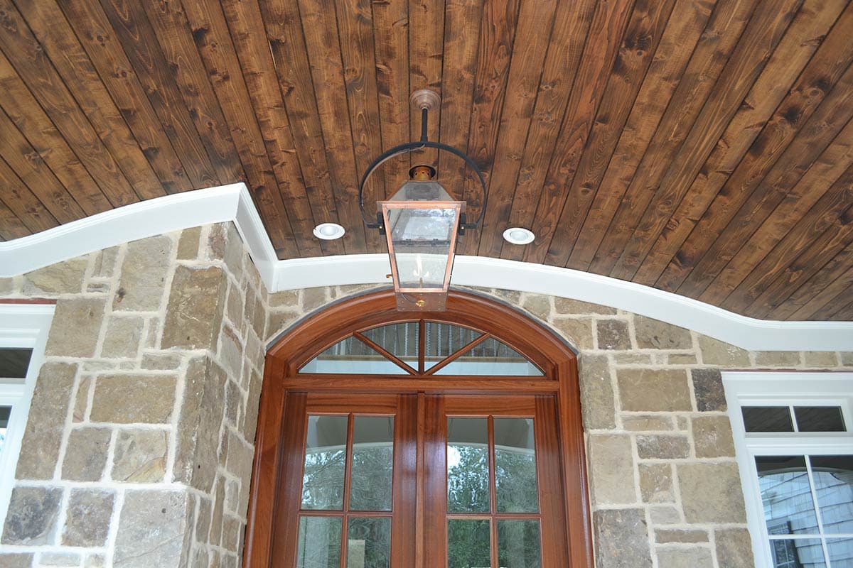 Exterior porch with a wooden ceiling, a decorative lantern, and stone walls. An arched wooden door is centered below the lantern.