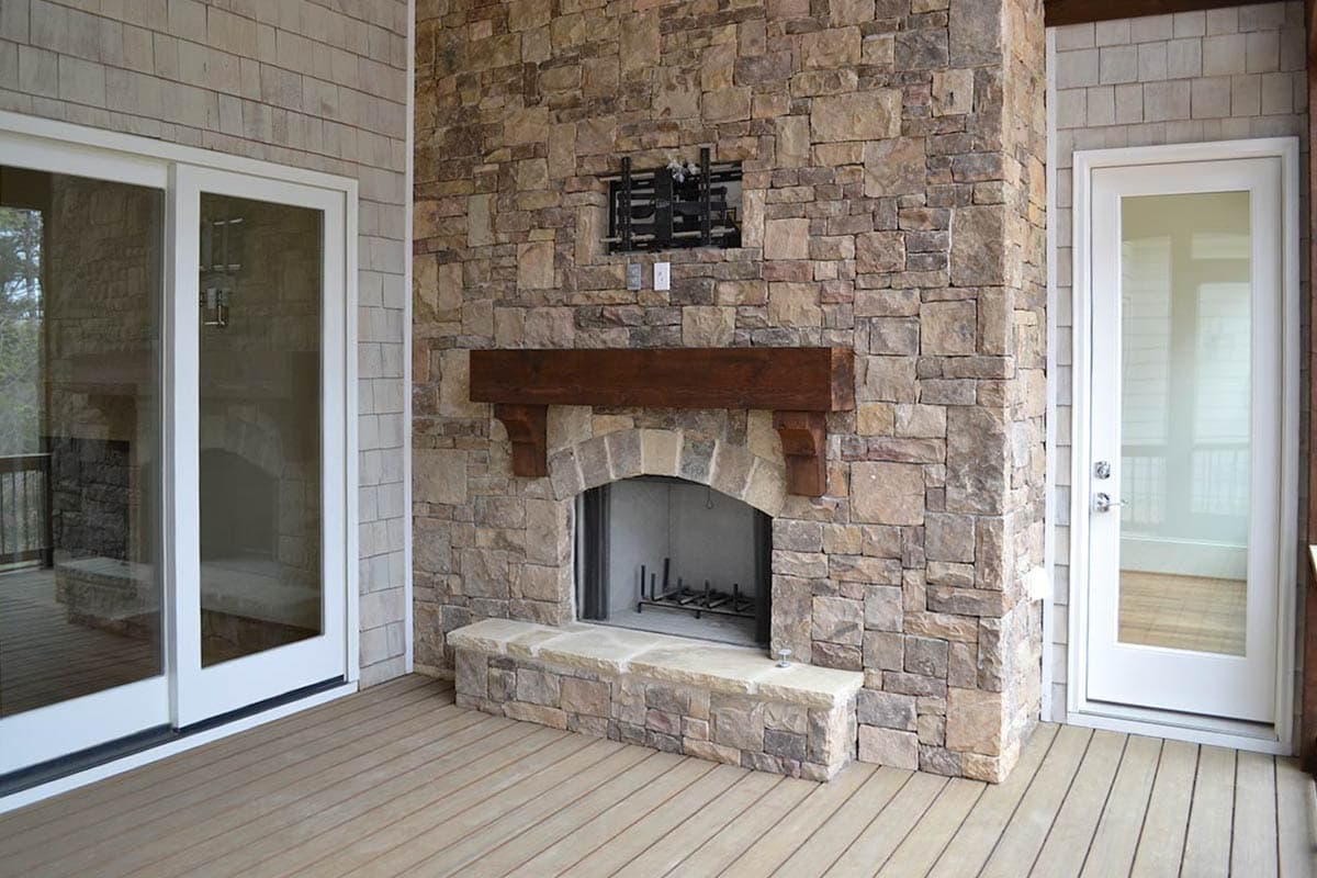 A stone fireplace with a wooden mantle sits on a porch, next to glass doors and a white-framed door. The floor is wooden planks.