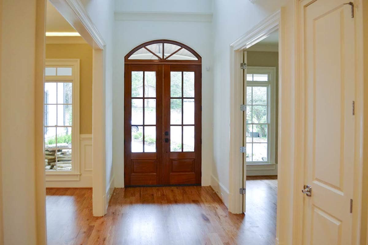 A sunlit hallway leads to a dark wood double door with glass panes. Hardwood floors and off-white walls create a welcoming atmosphere.