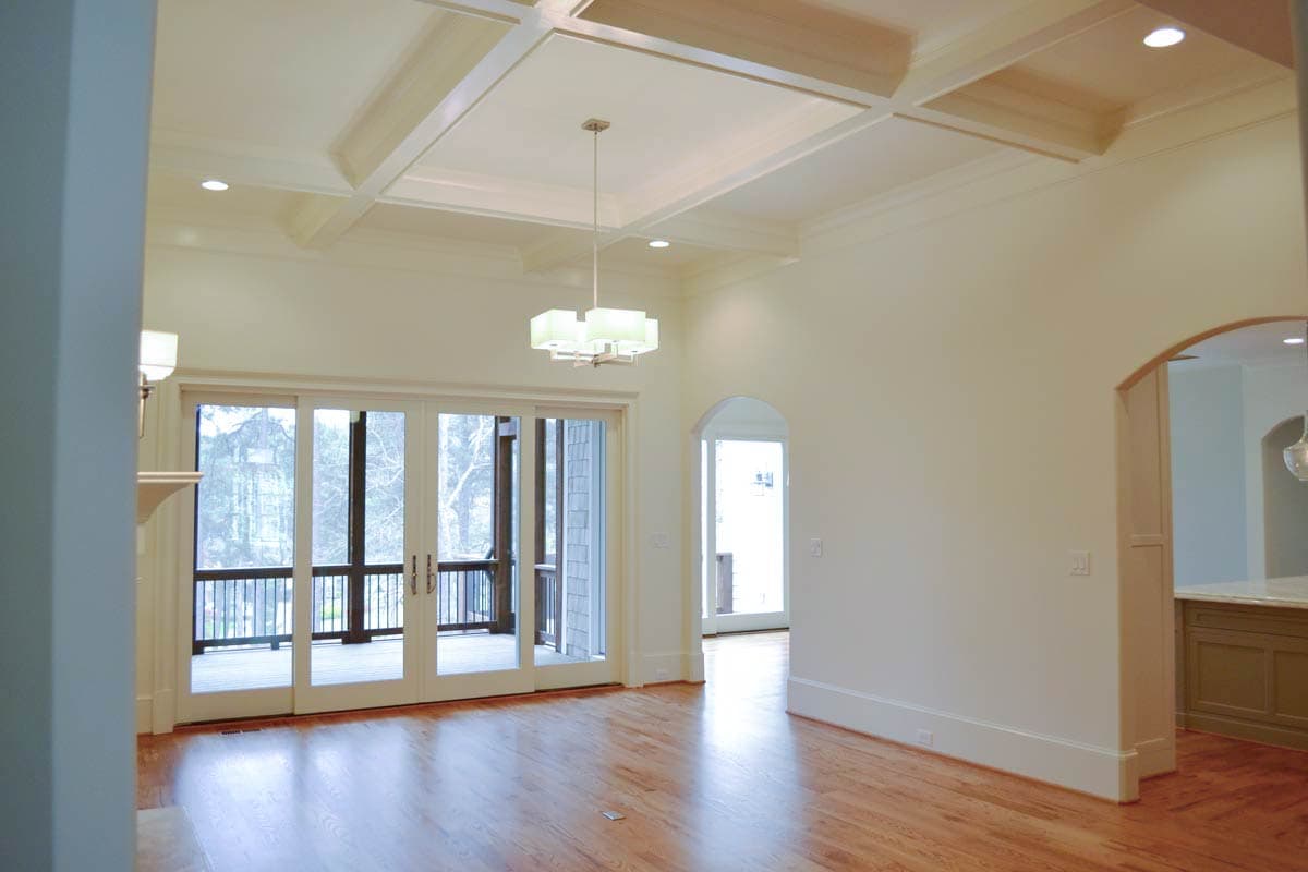 Bright, empty room with a coffered ceiling and hardwood floors. Glass doors lead to a porch and a small archway to another area.