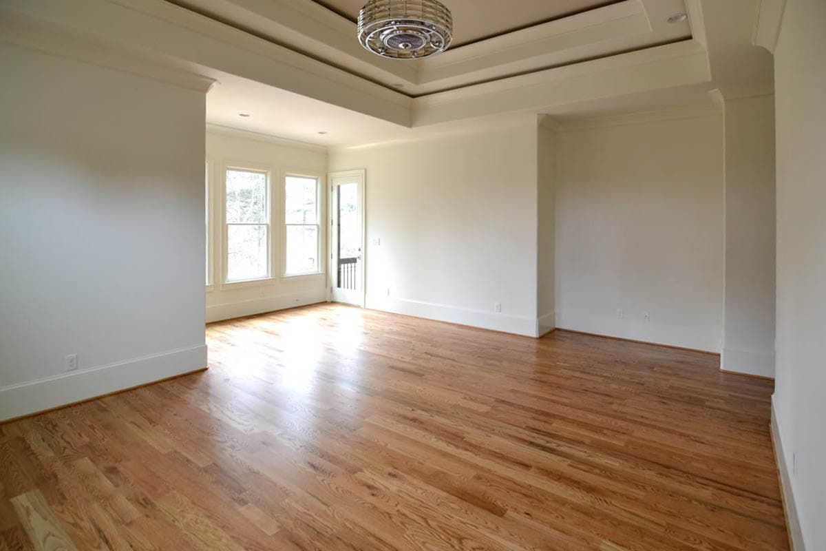 An empty, sunlit room with hardwood floors, white walls, and a recessed ceiling with a light fixture. Three windows and a door are visible.