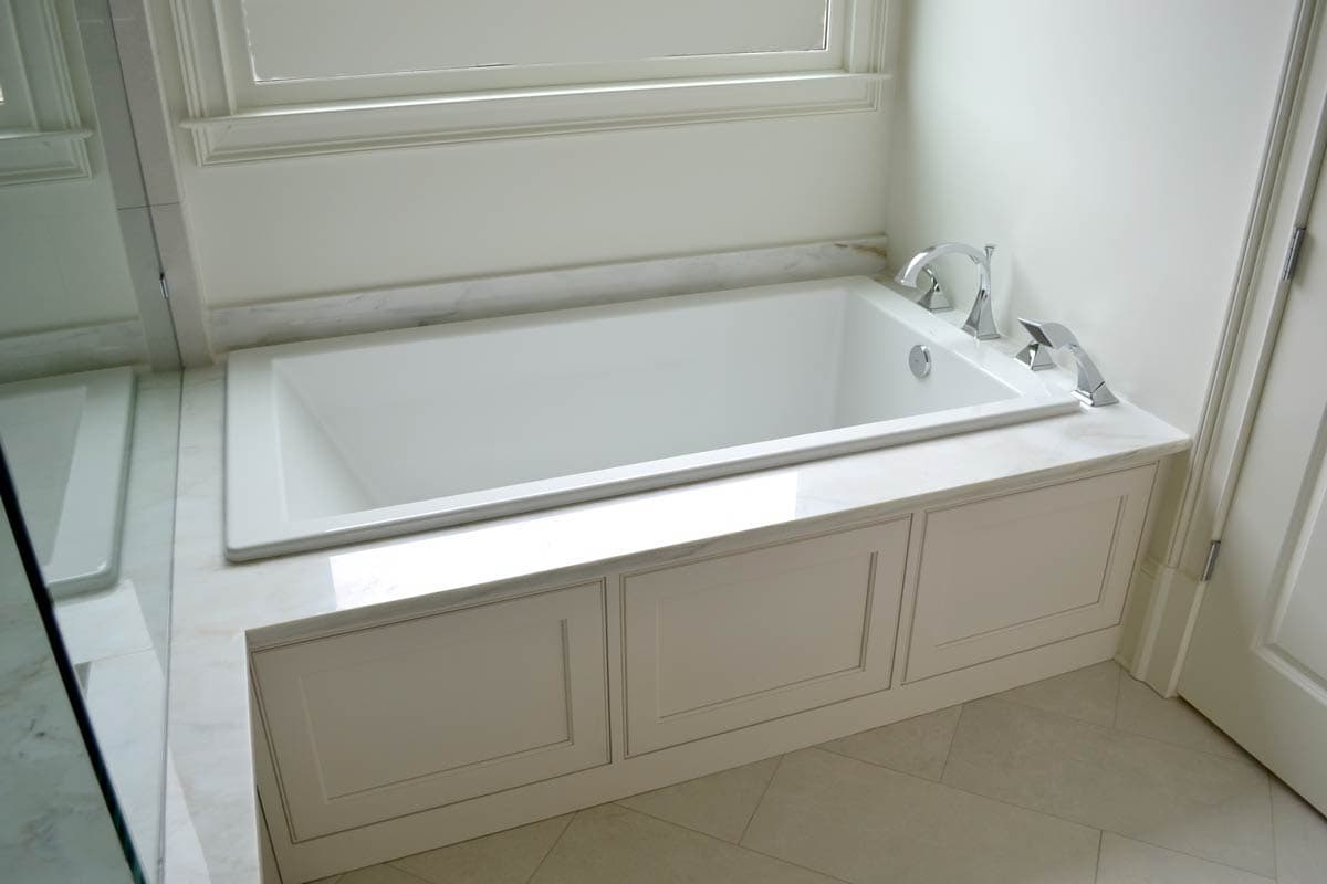A built-in white bathtub with chrome fixtures sits against a window in a bright bathroom. Decorative paneling lines the tub's base.