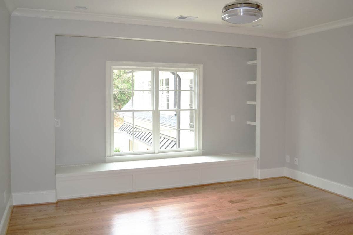 A light-filled room features a large window with a built-in window seat. Wooden floors and neutral walls create a spacious and modern aesthetic.