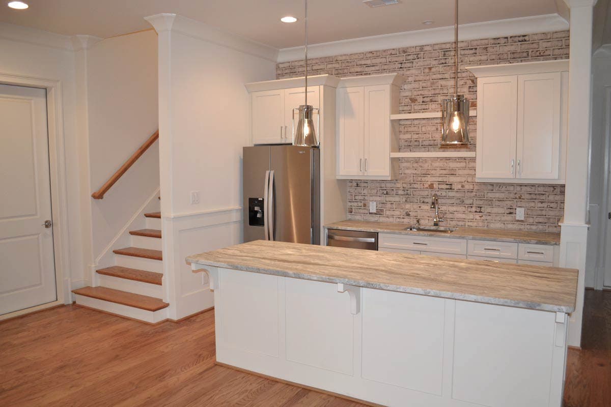 A modern kitchen with a large island, stainless steel appliances, and a brick backsplash. Stairs lead up on the left.