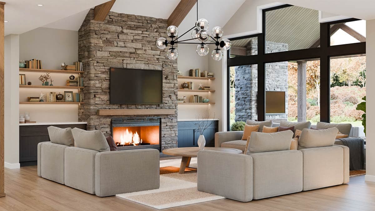 Cozy living room with a stone fireplace, lit fire, and two neutral-toned sofas. Large windows frame an outdoor view, and a modern chandelier hangs.