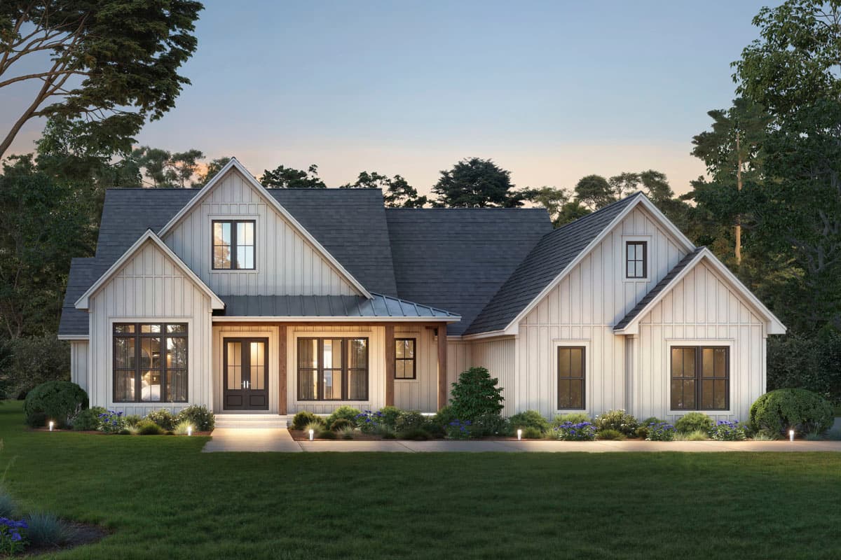 A white farmhouse with a dark gray roof, set at dusk. Exterior lights illuminate the path leading to the front door, surrounded by lush landscaping.
