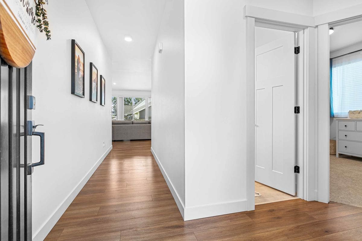 Interior hallway with hardwood floors, white walls, and a modern door. Living room visible in background.