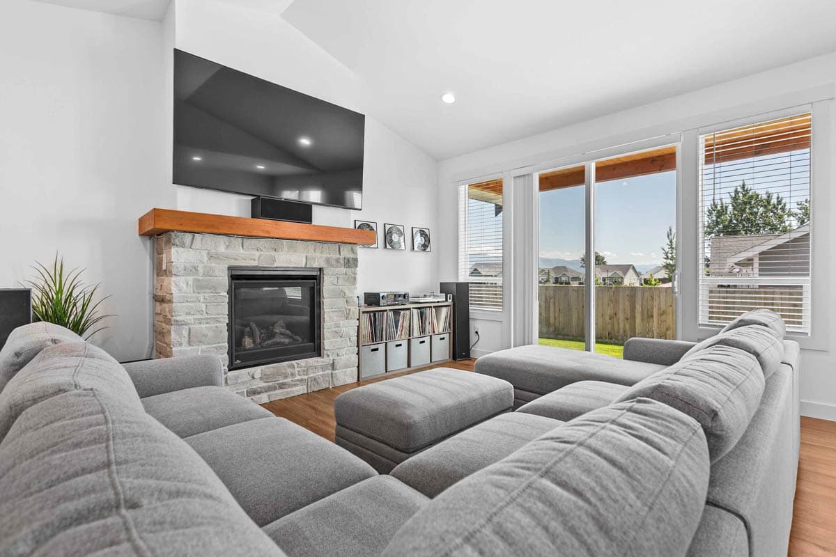 Interior view of a living room with a stone fireplace, mounted television, sectional sofa, and large windows overlooking a backyard.