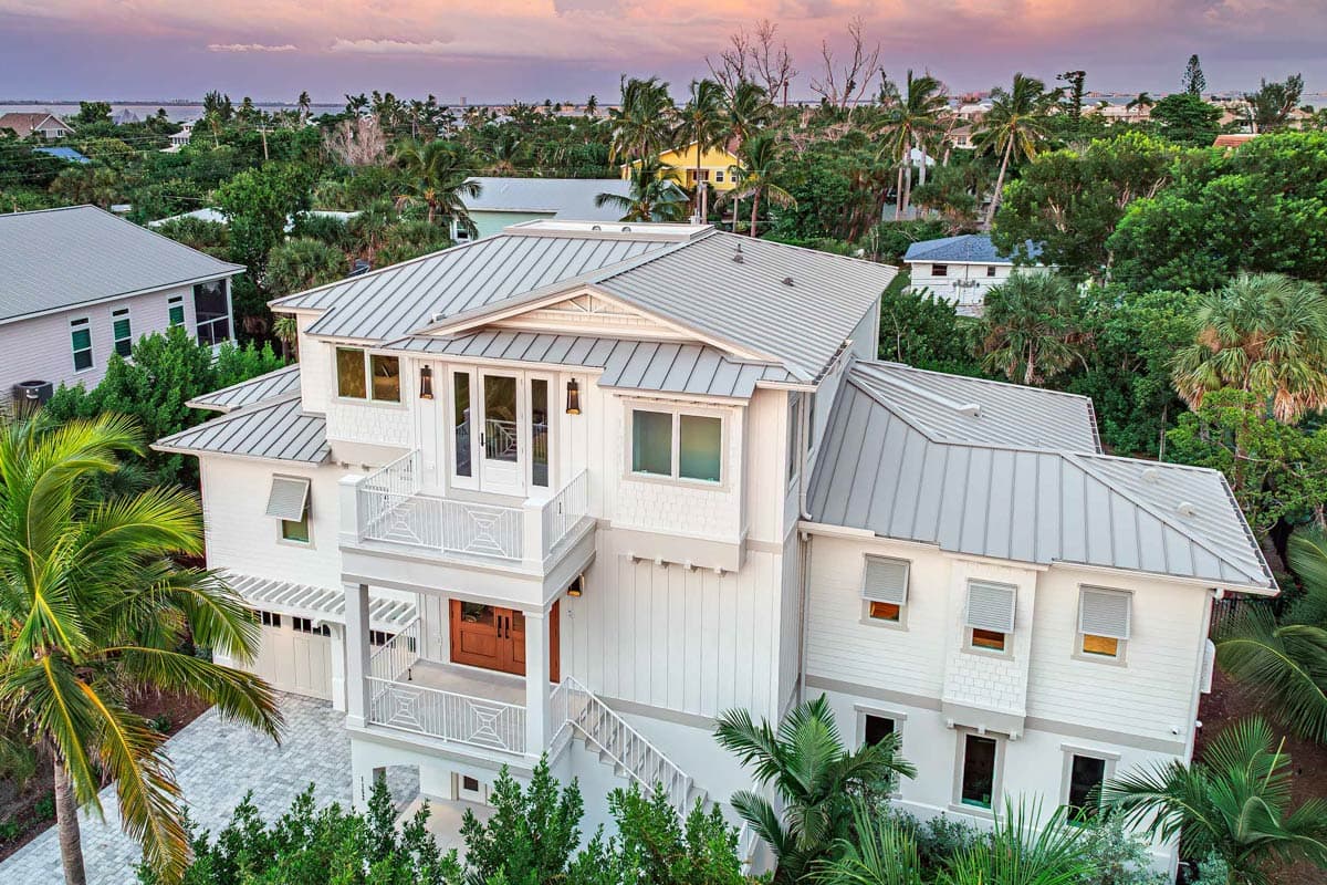 A multi-story, white coastal home with a metal roof and balconies. Lush green trees surround the house, with a sunset sky in the background.