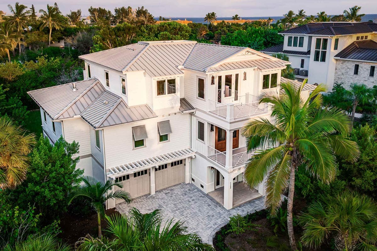 An aerial view showcases a luxurious white multi-story house with a gray metal roof, surrounded by lush tropical vegetation.