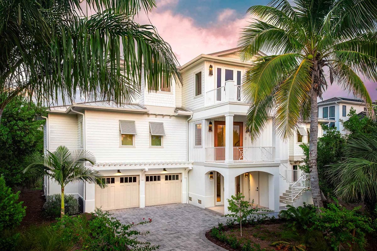 A white, multi-story house with a balcony and a driveway, surrounded by palm trees under a colorful sky at dusk.