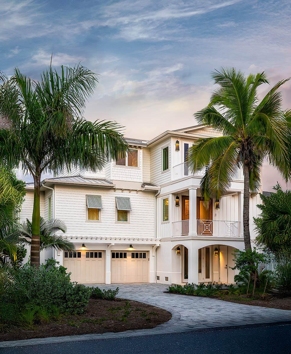 A large white coastal home with multiple levels, two-car garage, and a balcony. Palm trees frame the driveway and building.