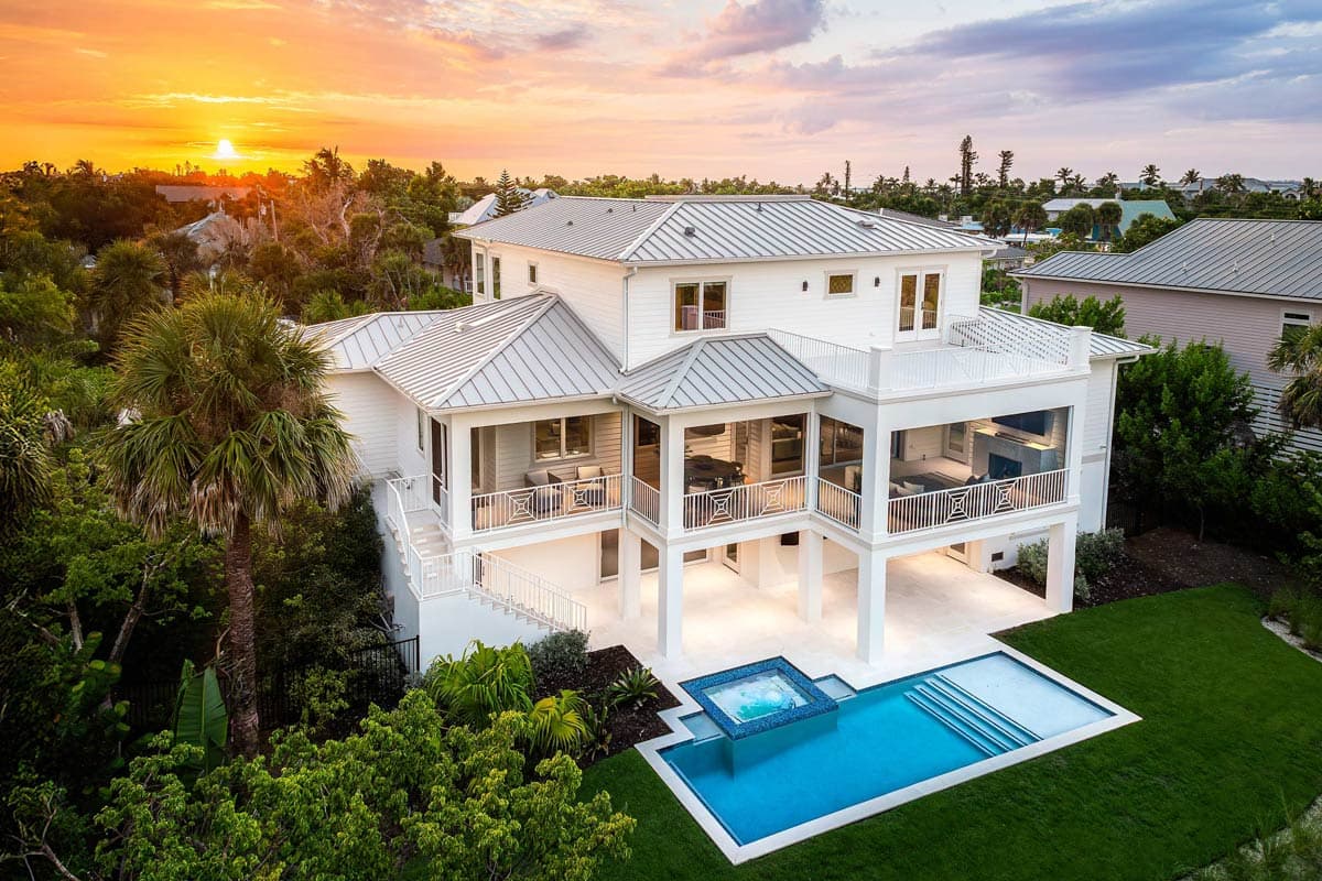 A modern, white coastal home with a pool and ocean view at sunset. Features include multiple balconies and lush landscaping.
