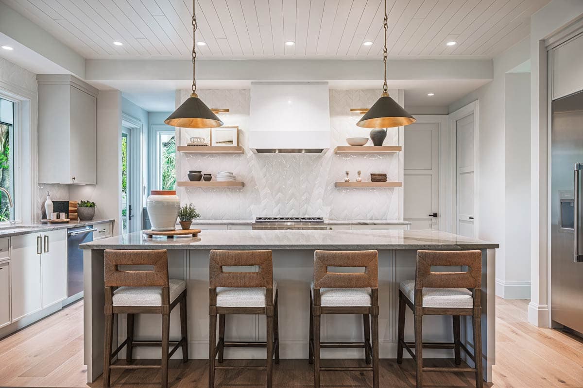 Stylish kitchen with an island, four bar stools, and pendant lights. White cabinets, herringbone backsplash, and open shelves are visible.