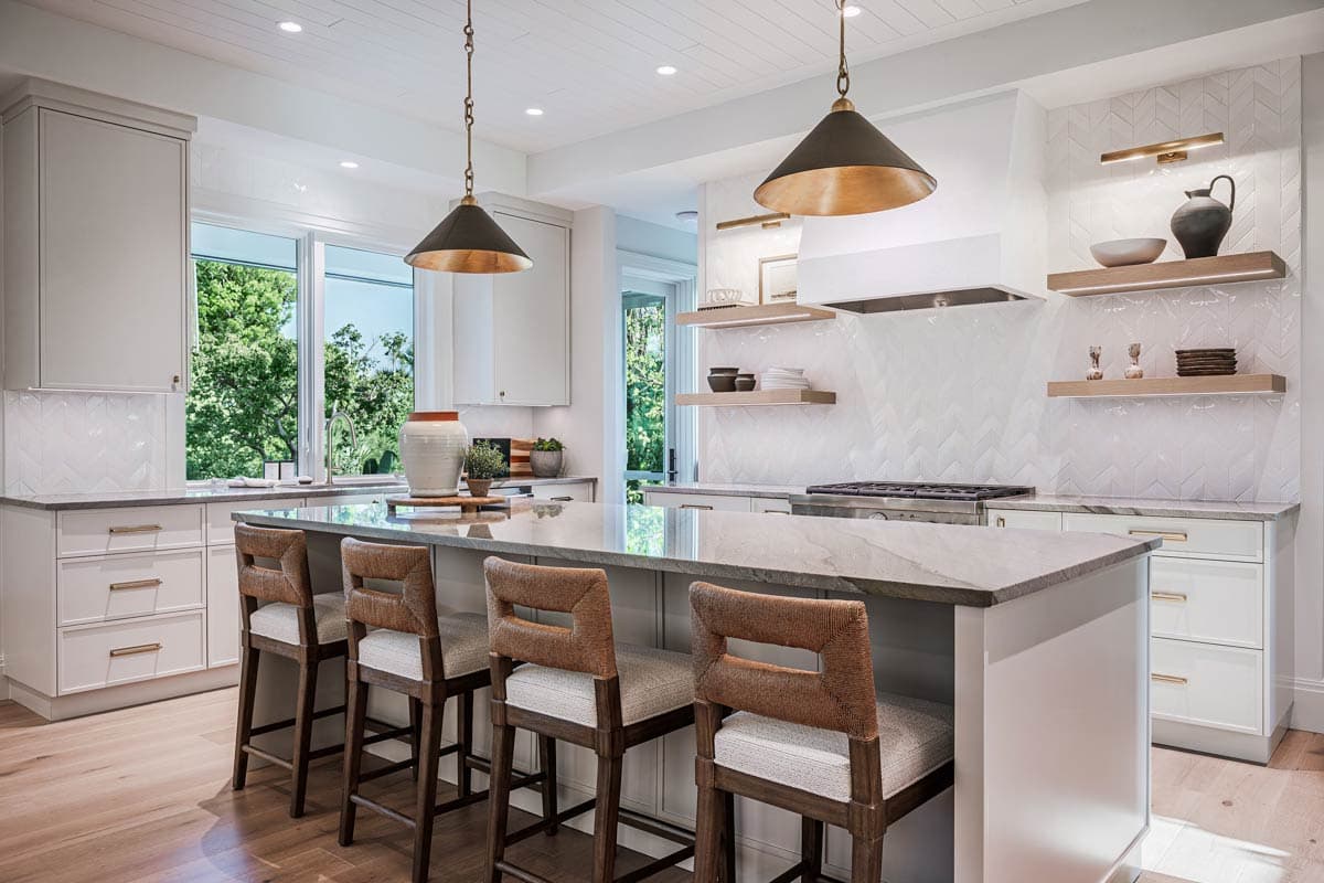 A modern kitchen with a large island and bar seating. Two pendant lights hang over the island, and a window shows the trees outside.