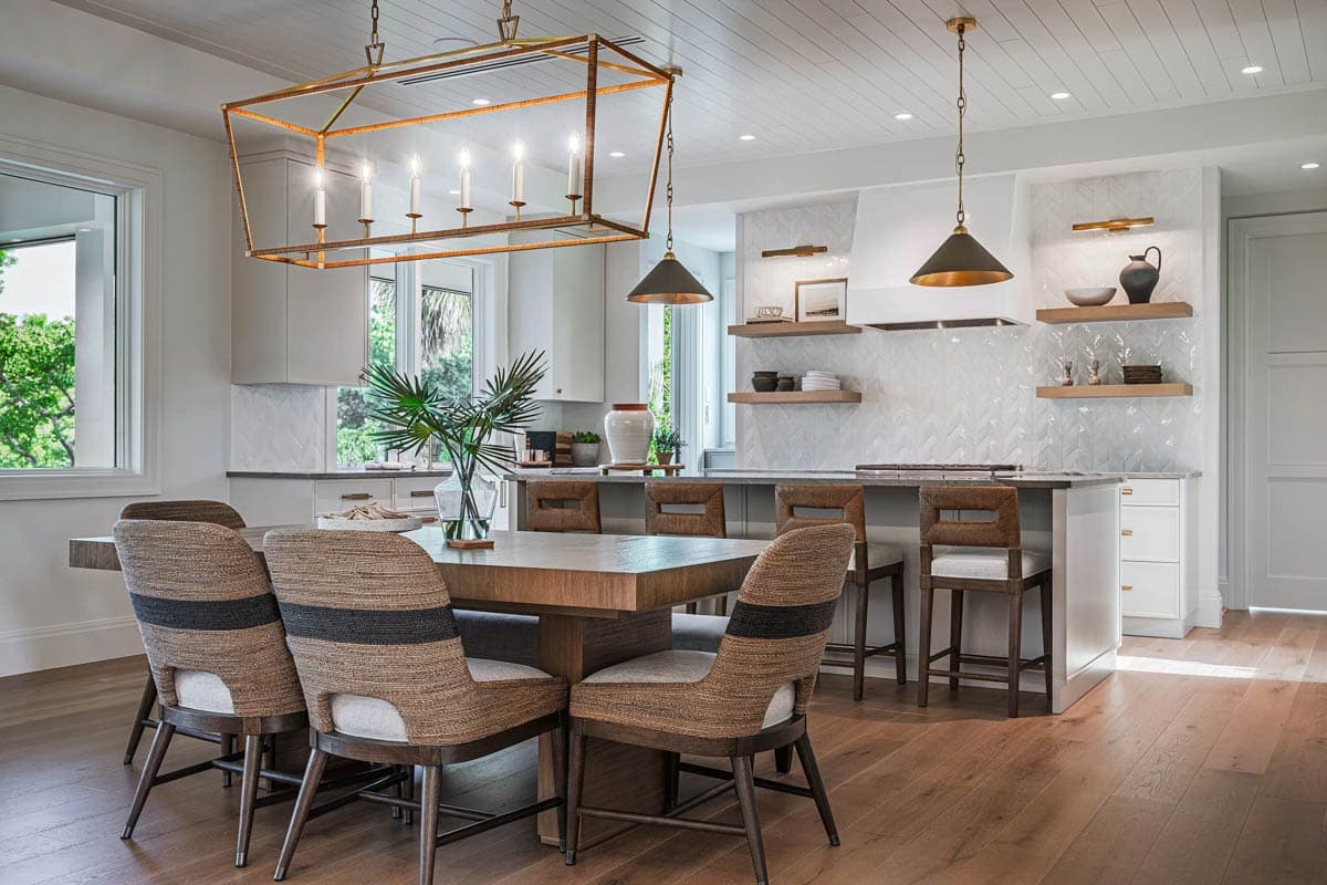 A modern dining room features a large rectangular table with stylish chairs. Overhead, a rectangular chandelier hangs. Further in, a kitchen area.