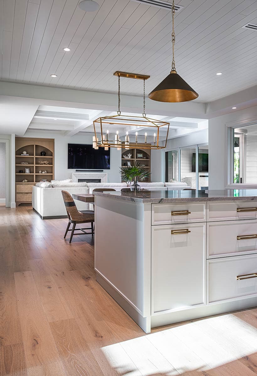 A modern kitchen and living room open to each other. White cabinets with gold hardware are in focus, with a large chandelier overhead.