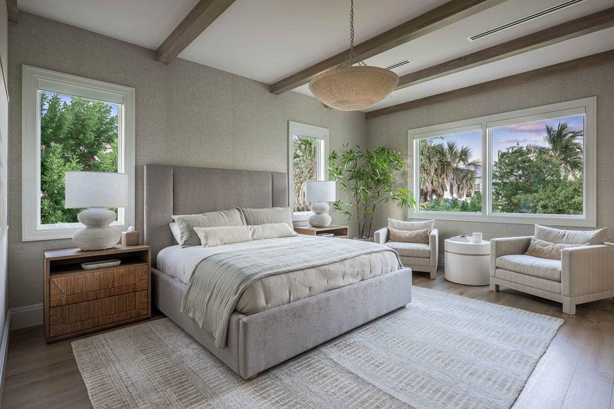 A luxurious bedroom features a large upholstered bed, neutral tones, and abundant natural light from large windows overlooking greenery.